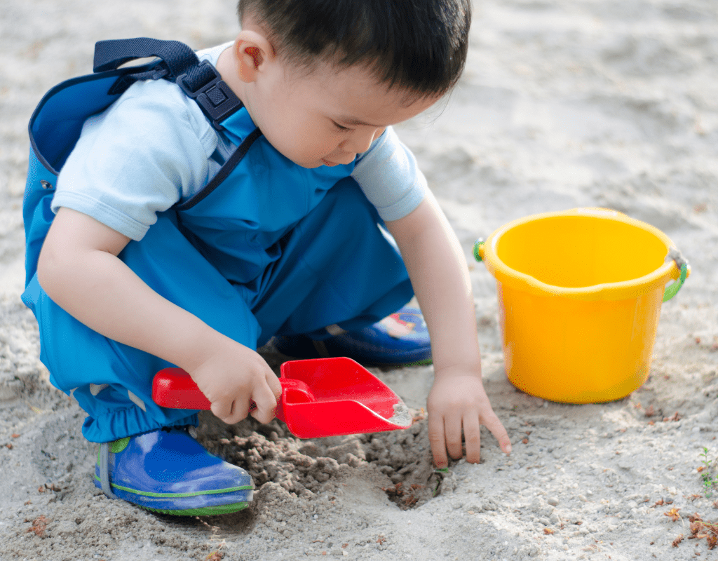 young child in blue pants playing with sand box toys in a sand box with a yellow bucket and red shovel